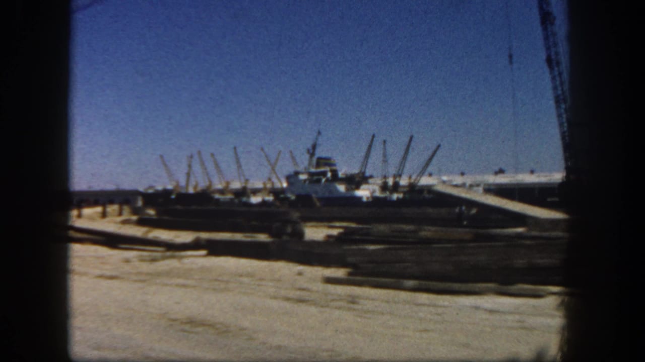 Construction site with a crane, scattered materials, and a vessel-like form beyond a dirt lot.