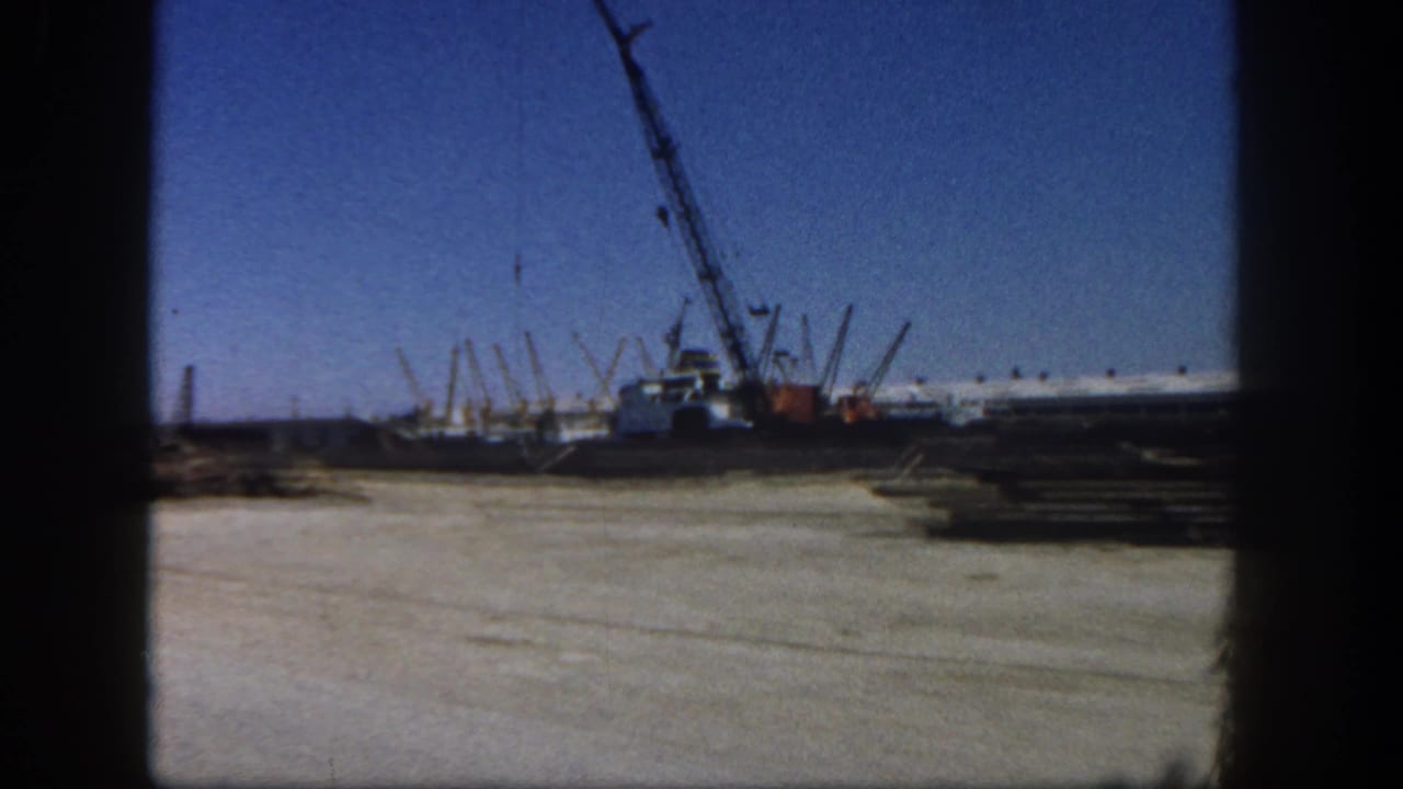 1962 San Diego 8mm frame showing a crane, worksite ground, and stacked lumber near boat docks under a clear sky.