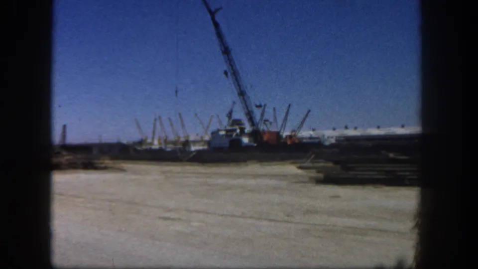1962 San Diego 8mm frame showing a crane, worksite ground, and stacked lumber near boat docks under a clear sky.