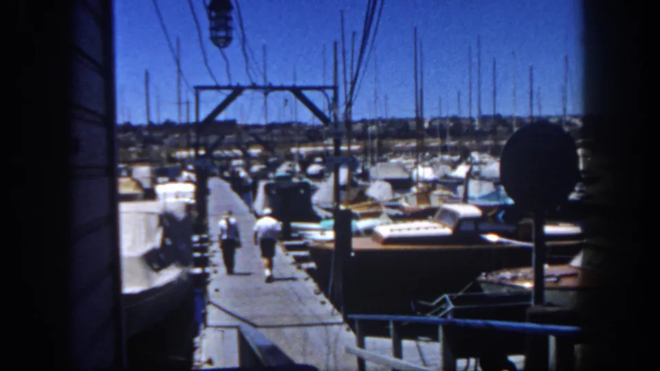 A 1962 San Diego marina frame shows two adults on a dock among moored sailboats and dense masts under bright midday light.