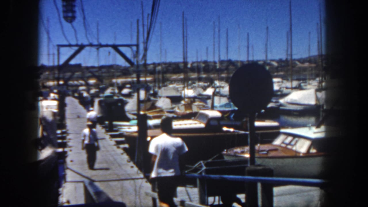In 1962 San Diego home-movie footage, a man runs down a dock past wooden boats and sailboat masts at a marina.