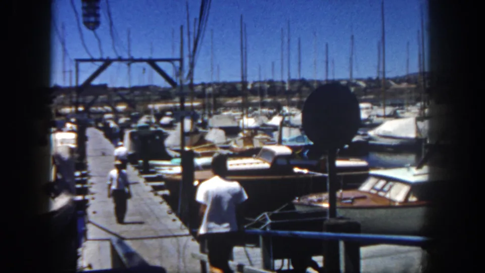 In 1962 San Diego home-movie footage, a man runs down a dock past wooden boats and sailboat masts at a marina.