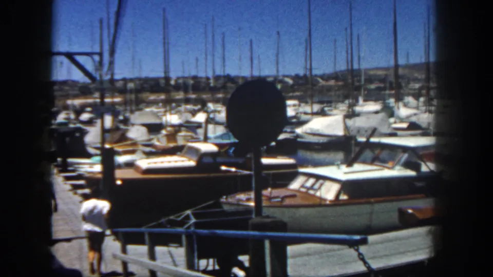 A man in light clothing is low on the dock near moored boats, with a crowded marina and masts in the background.