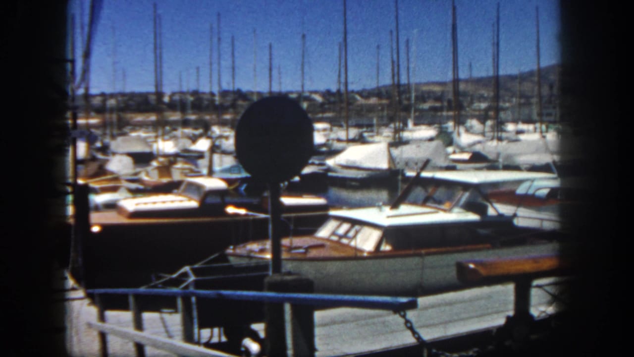 1962 San Diego marina view with docked cabin boats, sailboat masts, a round signpost, and dark foreground framing.