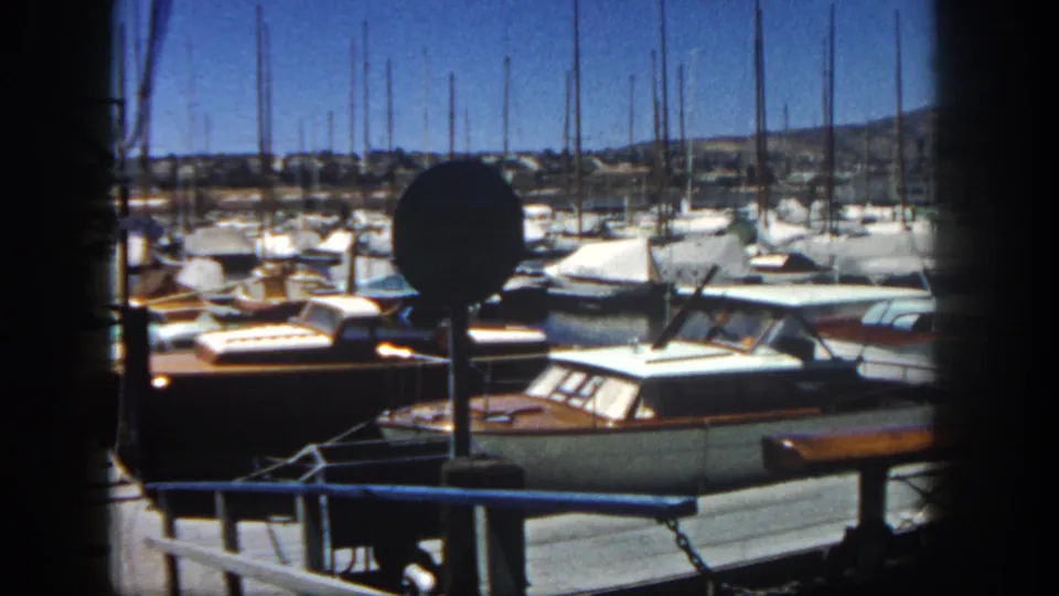 1962 San Diego marina view with docked cabin boats, sailboat masts, a round signpost, and dark foreground framing.