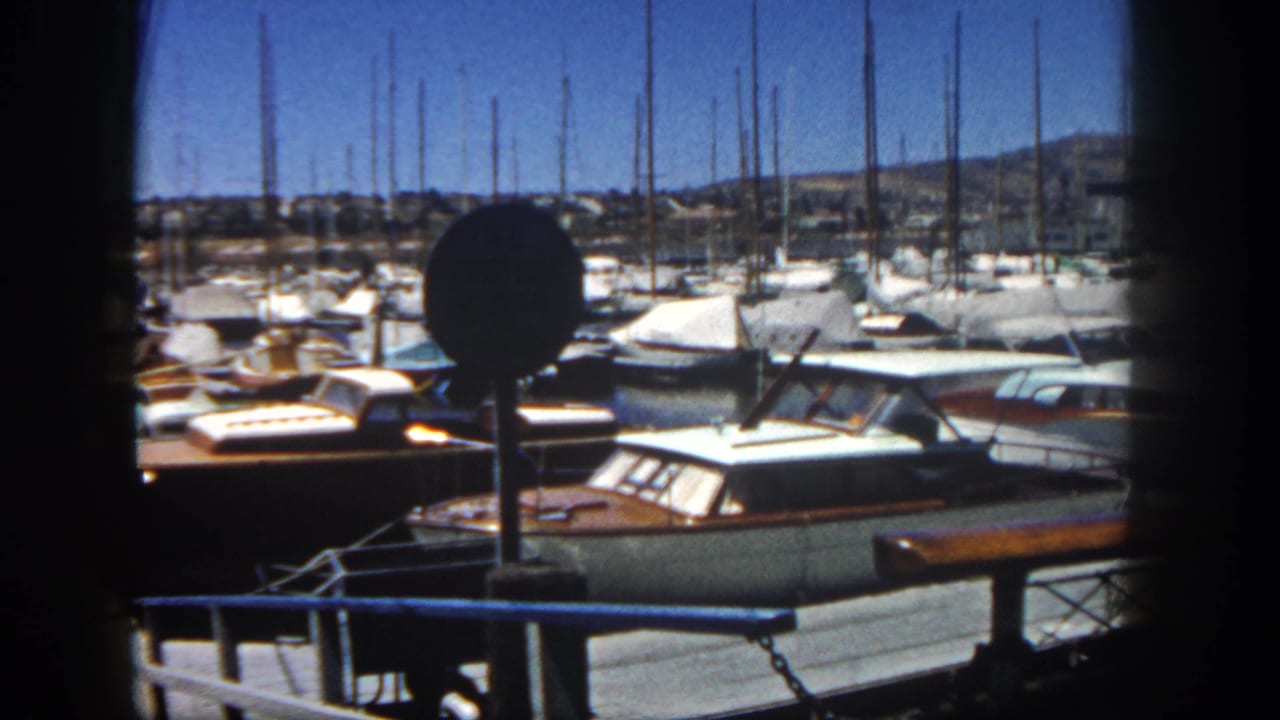Supported San Diego 1962 marina frame showing moored boats, sailboat masts, a dock railing, and low hills beyond the harbor.