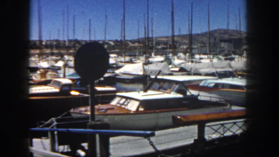 Home-movie frame of a 1962 San Diego marina, with classic docked boats, sailboat masts, railings, and a circular signpost.