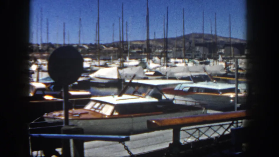 1962 San Diego marina still with docked boats in the foreground, dense sailboat masts, shoreline buildings, and low hills beyond.