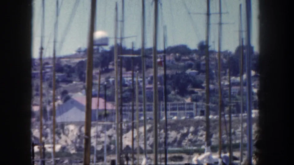 8mm view across San Diego marina masts toward hillside homes, shoreline buildings, and a water tower.