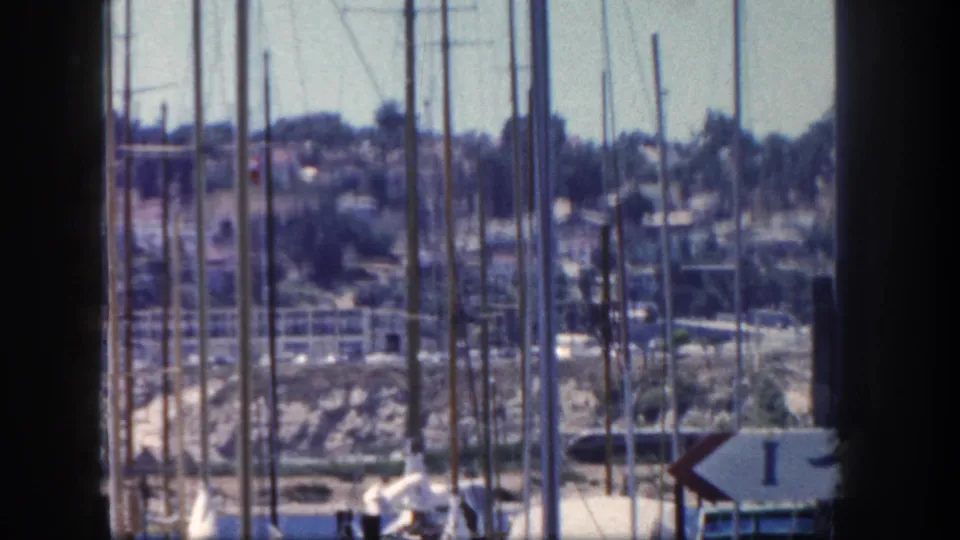Final 1962 San Diego marina frame with clustered sailboat masts, dock area, and hillside residences beyond.