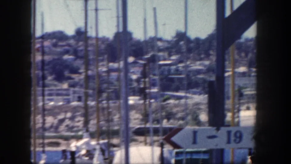 1962 San Diego marina view framed by dark structural posts, with sailboat masts, a hillside town, and a white I-19 sign in front.