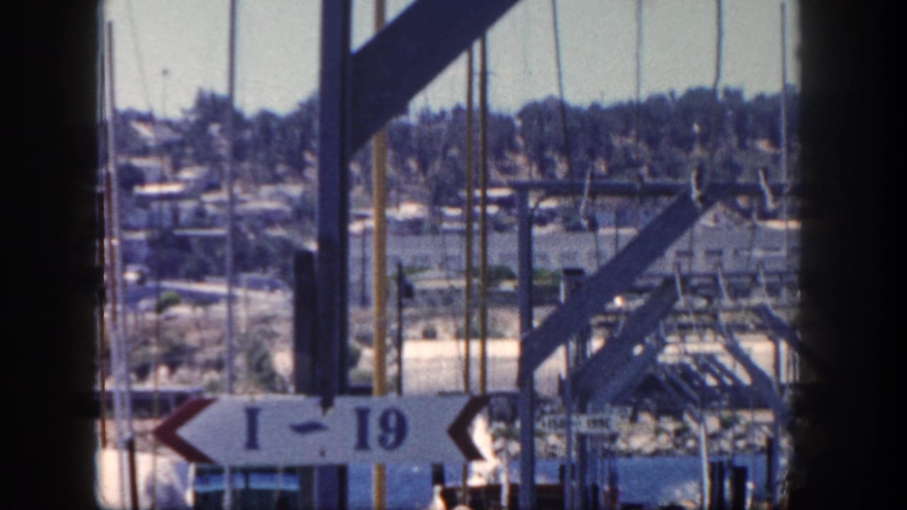 1962 San Diego harbor scene with sailboat masts, industrial-looking dock structures, power lines, hillside buildings, and an I-19 sign.