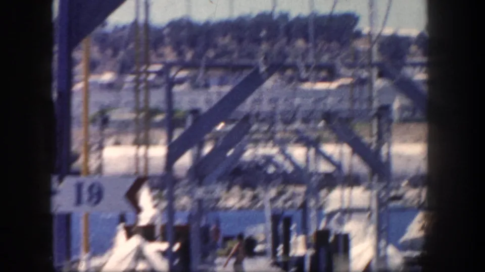 Blurred 1962 San Diego marina frame showing sailboat masts, dock structures, a partial 19 sign, dark posts, and distant hillside buildings.