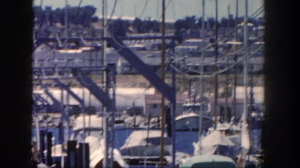 1962 San Diego marina view with clustered sailboat masts, angular dock framework, power lines, boats, water, and hillside buildings beyond.