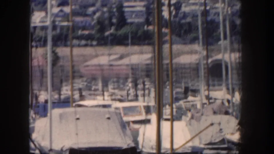1962 San Diego marina view with docked boats, tall masts, a dark foreground post, and hillside buildings in the distance.