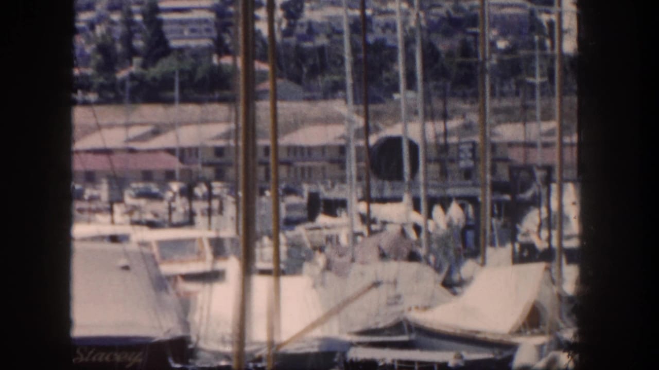 1962 San Diego marina view with sailboat masts, covered boats, dockside buildings, and a hilly background.