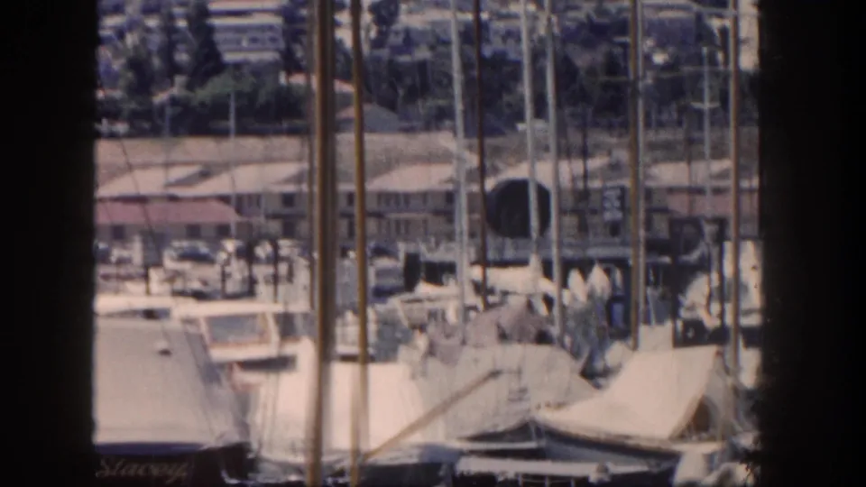1962 San Diego marina view with sailboat masts, covered boats, dockside buildings, and a hilly background.