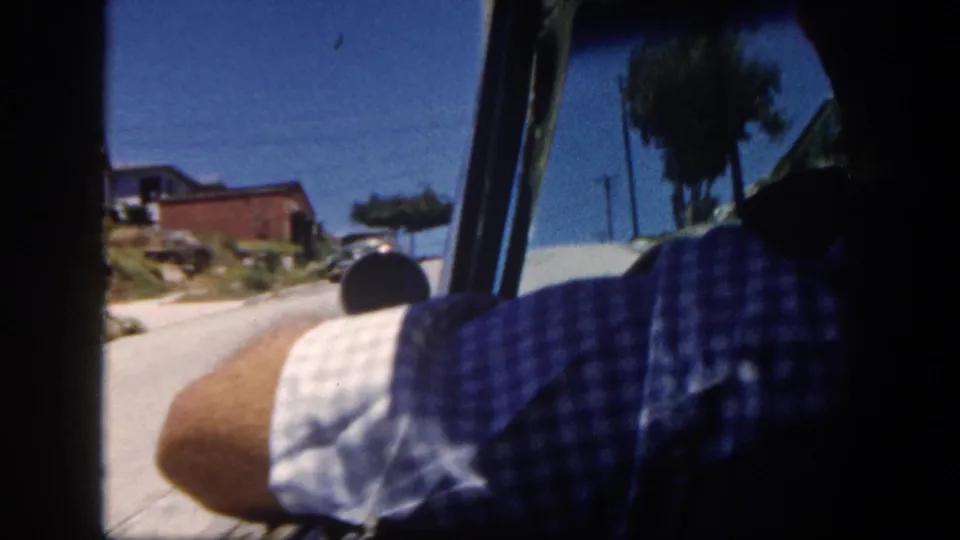 Inside-car view in 1962 San Diego, with a driver in a checkered shirt and a sunlit street ahead.