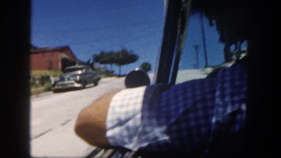 Passenger-seat view through a car windshield in 1962 San Diego, with a parked vintage car, utility poles, and low houses ahead.