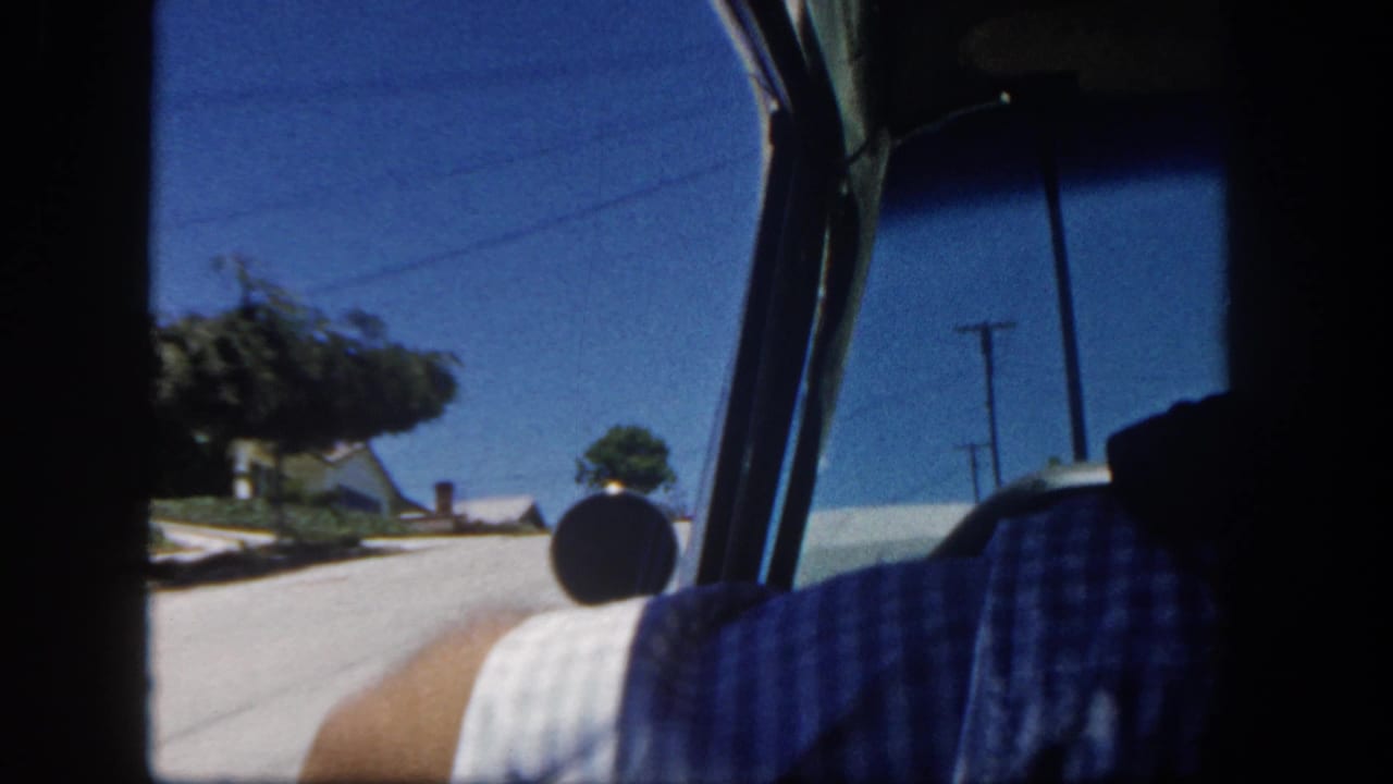 Interior car view in 1962 San Diego, with a partial passenger figure, residential street, houses, and utility poles visible.