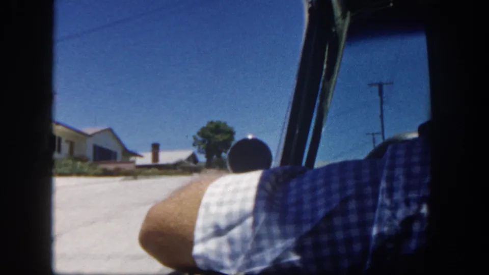 A 1962 San Diego residential driving view shows the car window, a passenger's arm, low houses, and a clear sky.