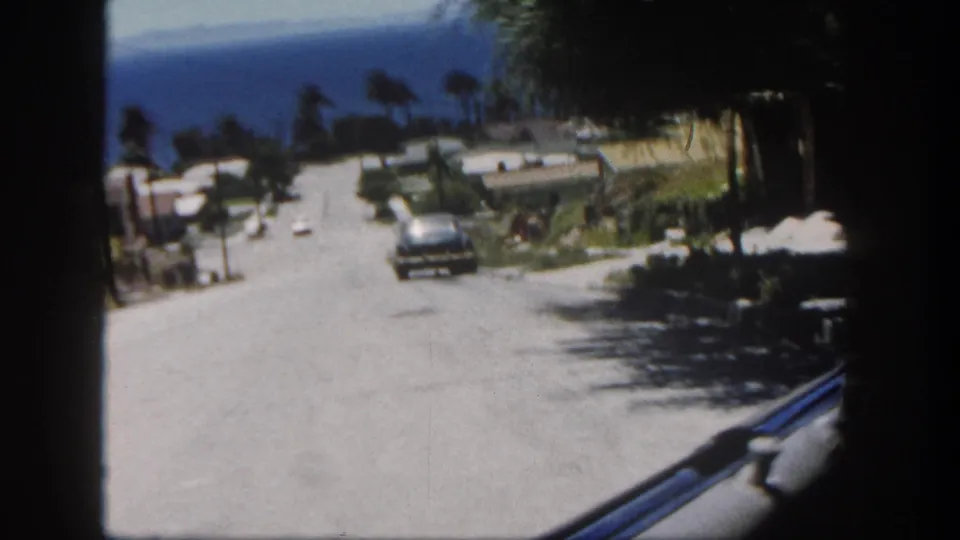 From inside a moving car in San Diego, the frame shows a residential street, parked cars, palms, and ocean in the distance.