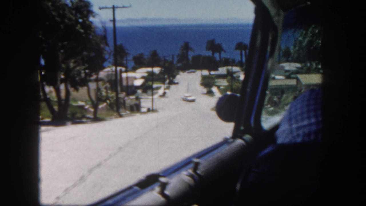 Downhill road view through a car window, with homes, trees, palms, and blue ocean beyond.