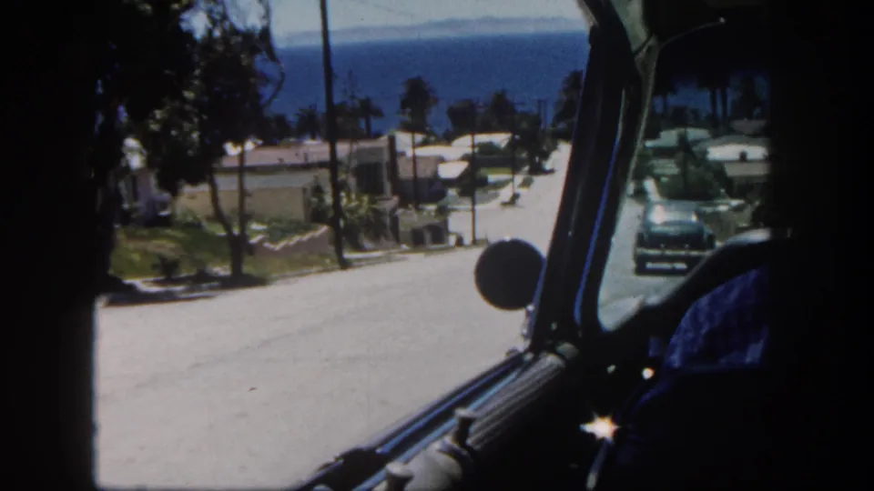 1962 San Diego driving view from inside a car, looking down a coastal hillside street as an oncoming automobile passes near homes and palms.