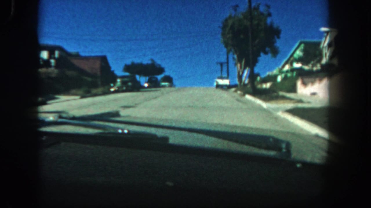 1962 San Diego windshield-view 8mm frame showing an uphill residential street, parked cars, houses, trees, and a bright blue sky.