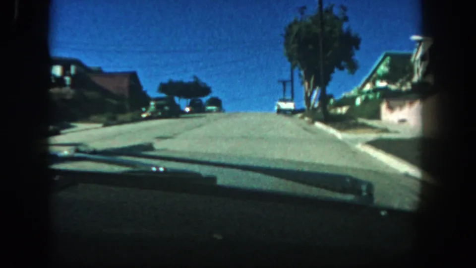 1962 San Diego windshield-view 8mm frame showing an uphill residential street, parked cars, houses, trees, and a bright blue sky.