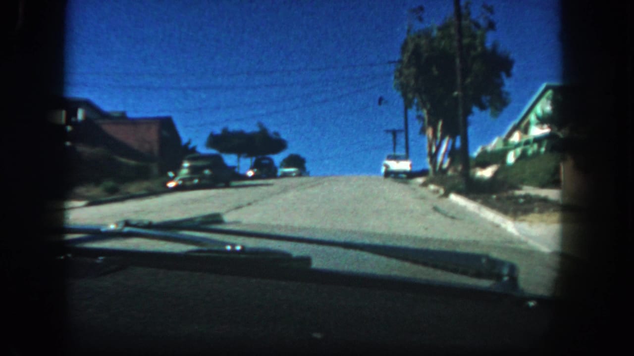 1962 San Diego home-movie frame from inside a moving car, looking uphill past parked cars, residential roofs, trees, and overhead lines.