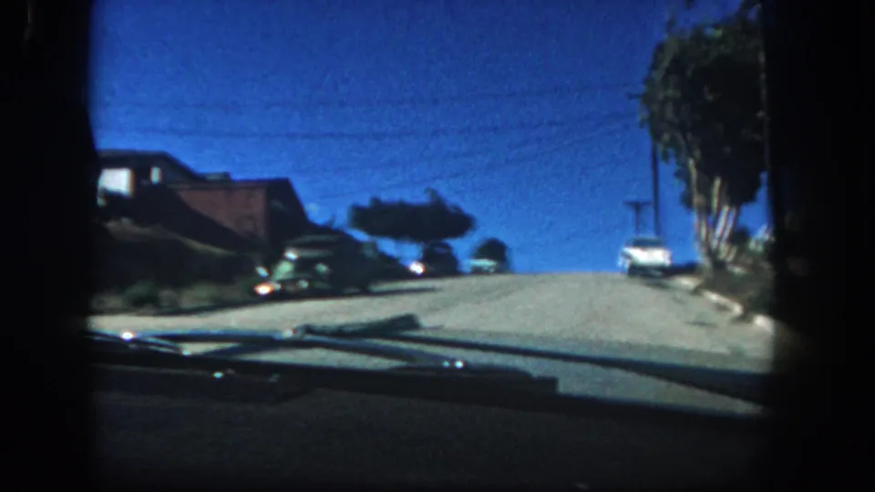 1962 San Diego 8mm frame showing a car moving uphill past curbside automobiles, modest houses, trees, and a saturated blue sky.