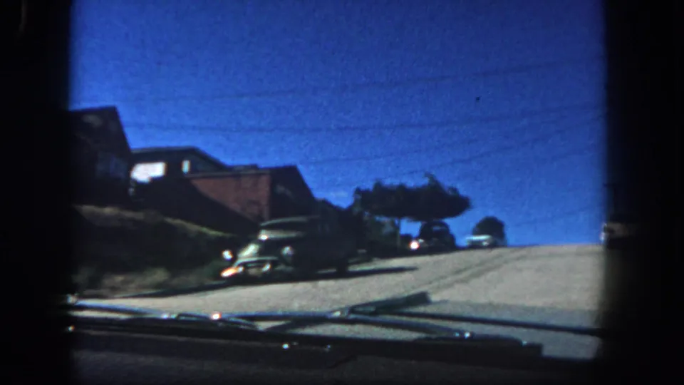 1962 San Diego driving frame looking toward the crest of a residential hill, with parked cars, low houses, trees, and dark 8mm frame borders.