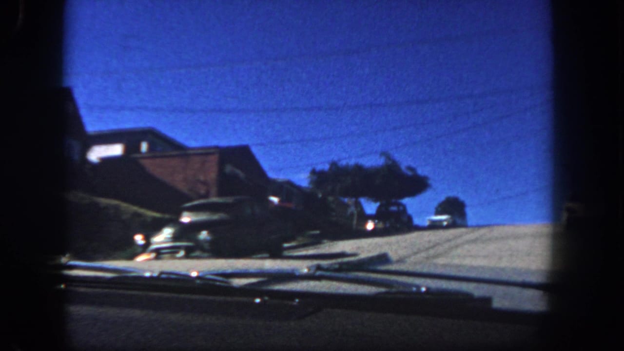 1962 San Diego windshield view showing a sloped residential street, parked mid-century cars, brick homes, trees, and the car dashboard.