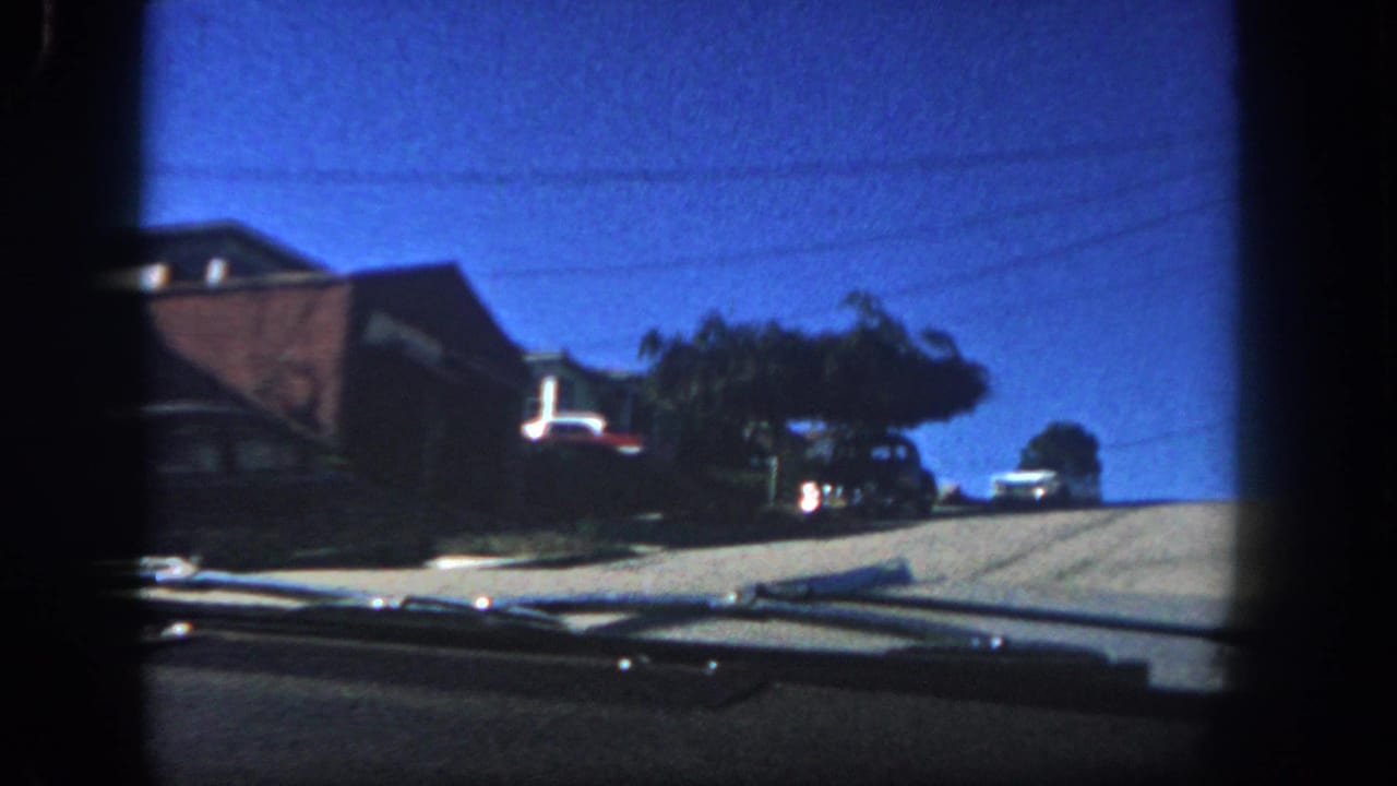 1962 San Diego car-window view with parked automobiles along a sloped suburban street, brick houses, trees, and dashboard edge.