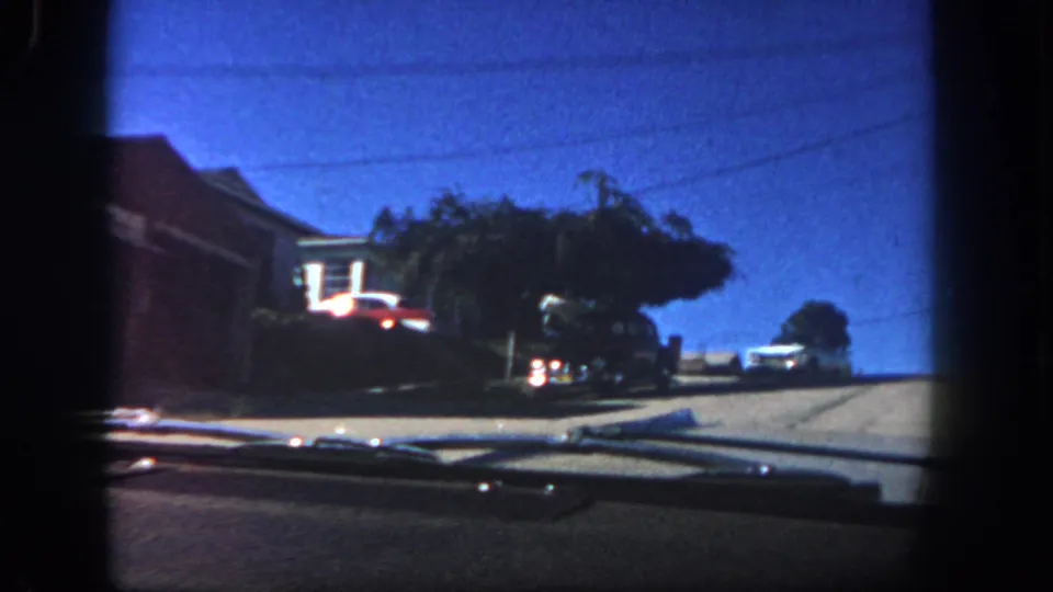 1962 San Diego windshield perspective showing a brick residence, parked cars, leafy trees, and an uphill neighborhood road.