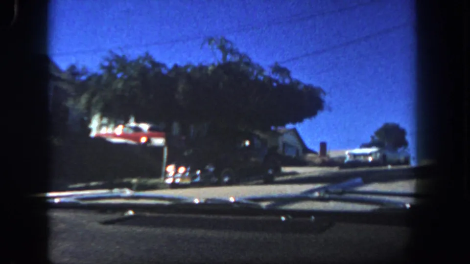 1962 San Diego driving view past trees and parked cars, with house fronts, dashboard trim, and a sunlit sloped street visible.