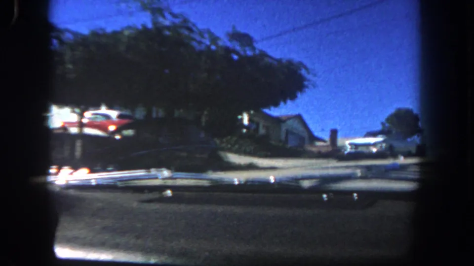 1962 San Diego home-movie frame showing a street, trees, parked cars, and rooftops through heavy dark film borders.