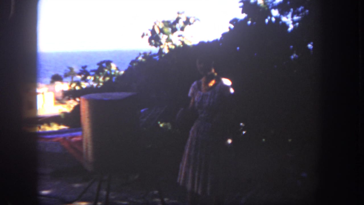 Woman in a dress standing in shadow on a patio with ocean visible beyond trees.