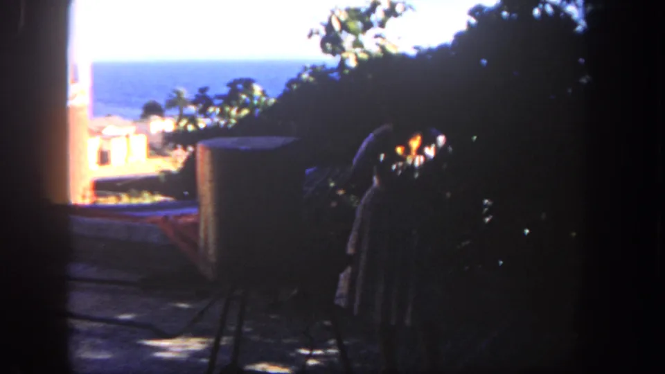 Home-movie frame from 1962 San Diego showing a woman on a shaded patio, foliage, rooftops, and blue ocean beyond.