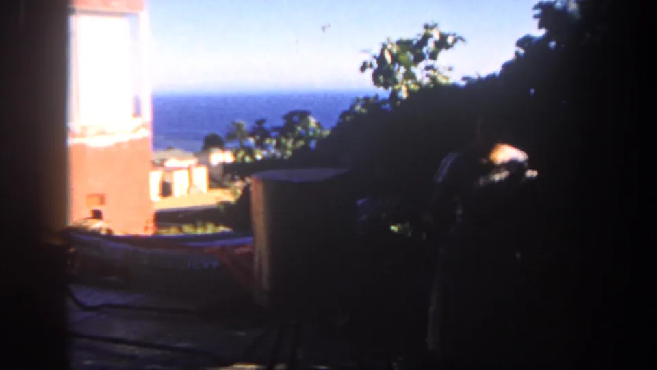 1962 San Diego 8mm frame with ocean horizon, patio wall, trees, rooftops, and a shadowed figure at the edge.