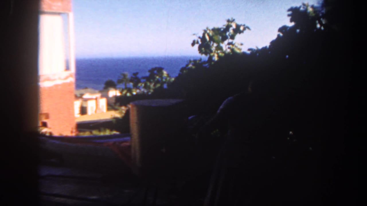 1962 San Diego 8mm frame looking from shade toward an ocean horizon, brick wall, patio furniture, foliage, and a partly hidden grill.