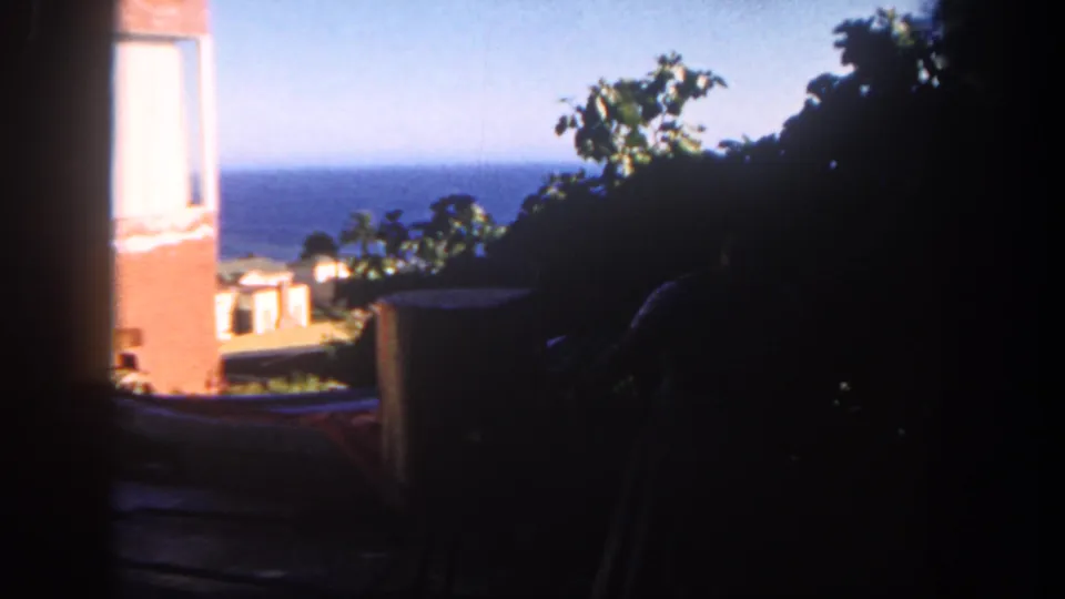 1962 San Diego 8mm frame looking from shade toward an ocean horizon, brick wall, patio furniture, foliage, and a partly hidden grill.