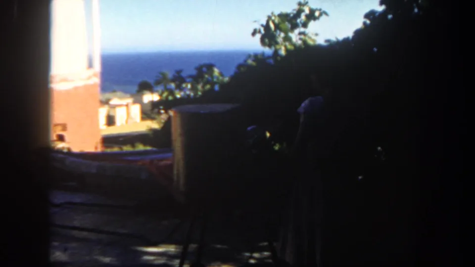 1962 San Diego 8mm frame of a shaded patio overlooking the ocean, with foliage, brickwork, and a grill form in deep shadow.