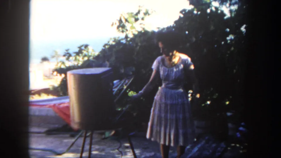 1962 San Diego home-movie frame showing a woman in a light patterned dress beside a patio grill, with foliage and ocean view behind.