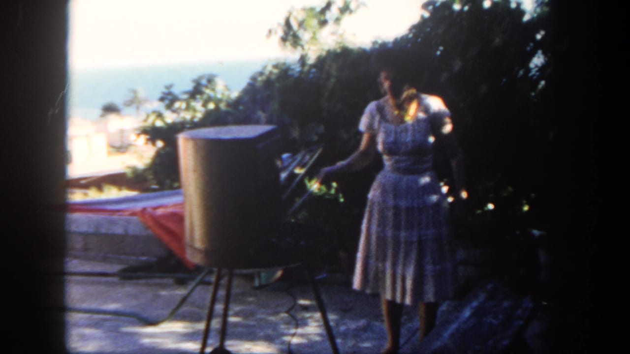 Woman in a patterned dress stands beside a cylindrical grill on an ocean-view patio.