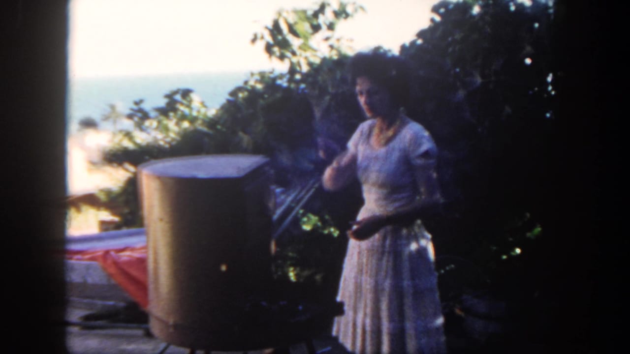 Woman gestures near a large patio grill with greenery and ocean in the background.