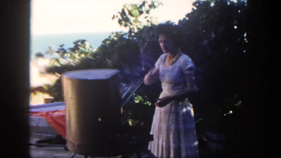 The woman stands near the cylindrical grill on a San Diego patio, framed by dark foliage and a bright ocean horizon.
