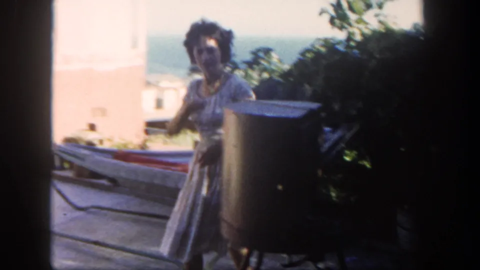 A woman in a light patterned dress stands by the grill, with patio furniture, foliage, and the Pacific visible in the background.