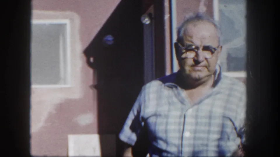 An older man in glasses and a light checked shirt stands near a doorway, with exterior windows visible behind him in the supplied 1962 San Diego footage.
